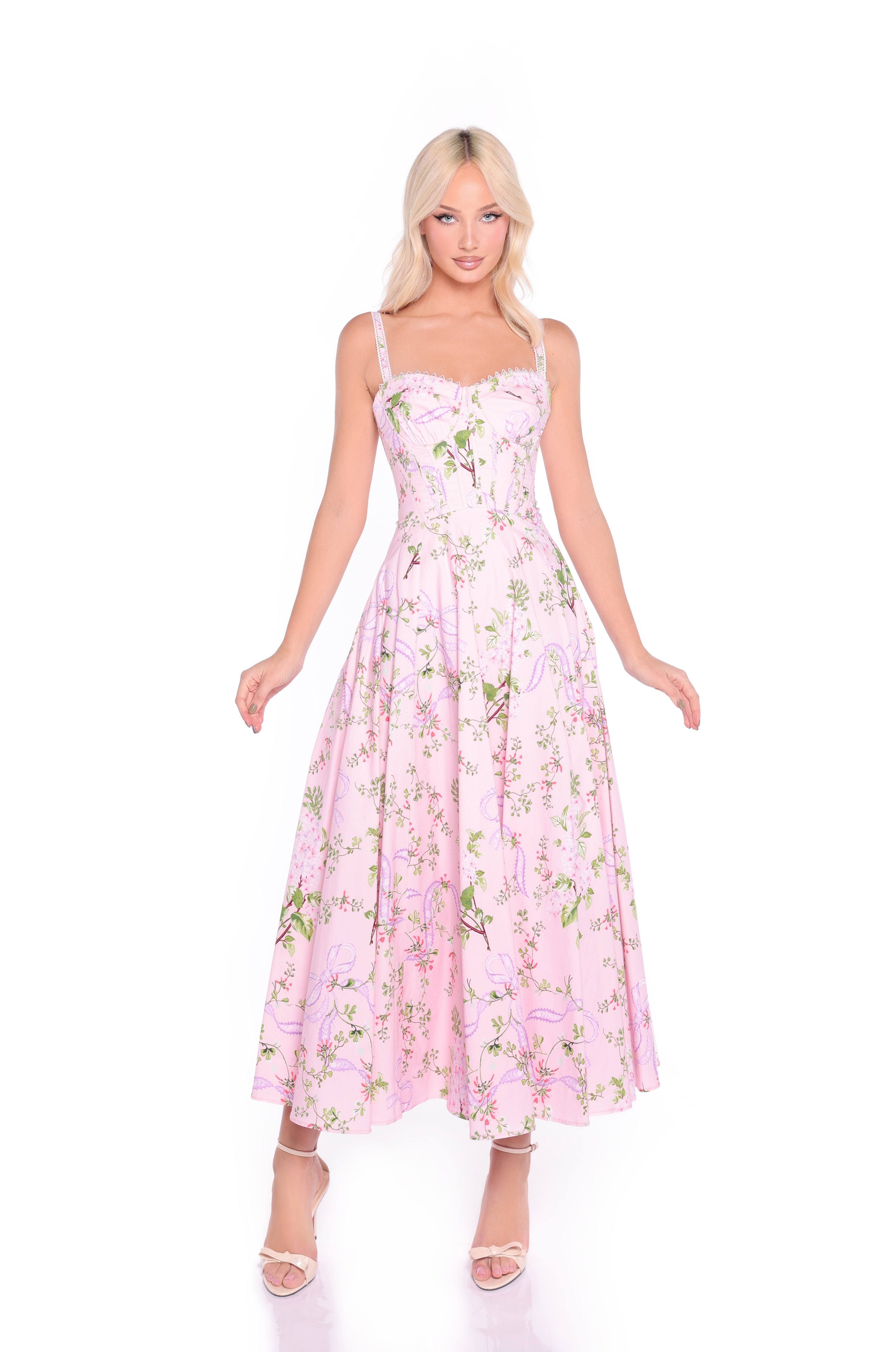 A woman wears the Rosella Blush Pink Floral & Bow Print Casual Dress, featuring a fitted bodice and flared skirt—perfect for summer weddings—paired with strappy heels, standing against a white background.